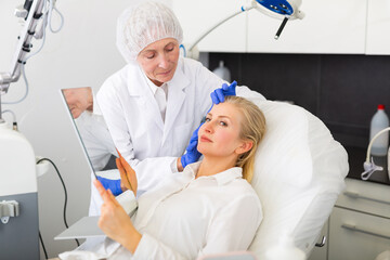 Experienced female cosmetologist giving professional advice before hardware facial skin treatments to adult woman looking at mirror in cosmetological office