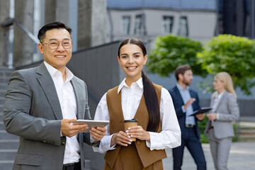 Two cheerful business colleagues with digital tablet standing outdoors, with other team members in the background.