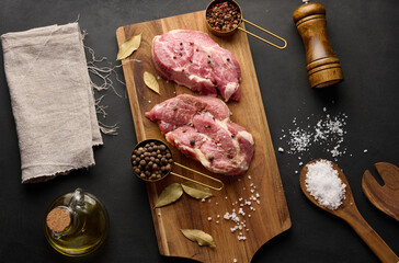 Wall Mural - Two raw pork neck steaks on a board and spices for cooking. Top view of black table.