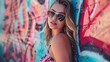 Beautiful young woman with long blond hair and blue eyes wearing sunglasses and a pink floral dress posing in front of a colorful graffiti wall.
