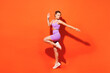 © ViDi Studio - Full body side view young fitness trainer instructor woman sportsman wearing purple top clothes train in home gym raise up leg hand isolated on plain orange background. Workout sport fit abs concept.