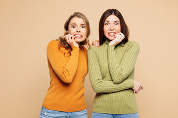 Young scared sad friends two women they wear orange green shirt casual clothes together look camera biting nails fingers isolated on plain pastel beige background studio portrait. Lifestyle concept.