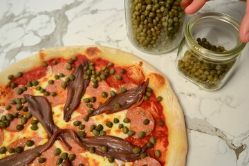 Poster - person arranging anchovies on pizza next to capers jar