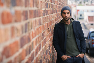 Poster - Portrait, confidence and man on brick wall in city in casual clothes, outfit or fashion for winter outdoor. Serious, cool and stylish person in urban town in beanie, hoodie and trendy jacket in Spain