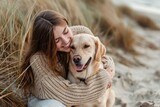 Fototapeta Zwierzęta - A young woman in a sweater hugs a golden retriever dog sitting on the sand near the sea