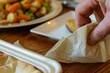 closeup of a hand unfolding a paper napkin with a meal in the background