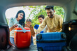 Indian couple with helping kids taking out luggage or picnic basket taking out from car boot - concept of destination, family travel and summer vacation or holidays