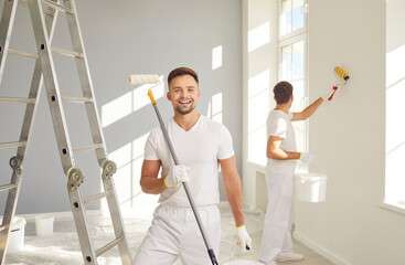 Wall Mural - Portrait of happy men professional painters painting a wall with paint roller and looking at camera. Building contractors doing repair renovation in client house. Repair and renovating concept.