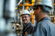 Two men wearing hard hats and safety glasses are smiling at the camera. They are likely workers in a factory or industrial setting