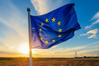 The blue flag of the European Union with twelve yellow stars flutters against a sunrise background