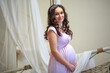 Portrait of a beautiful young pregnant woman in a pink dress. A beautiful pregnant woman in a bright room hugs her stomach with her hands