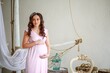 Portrait of a beautiful young brunette woman in a pink dress. A beautiful pregnant woman in a bright room hugs her stomach with her hands
