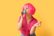 Beautiful young woman in pink wig with blue sweet macaroon showing heart gesture on yellow background