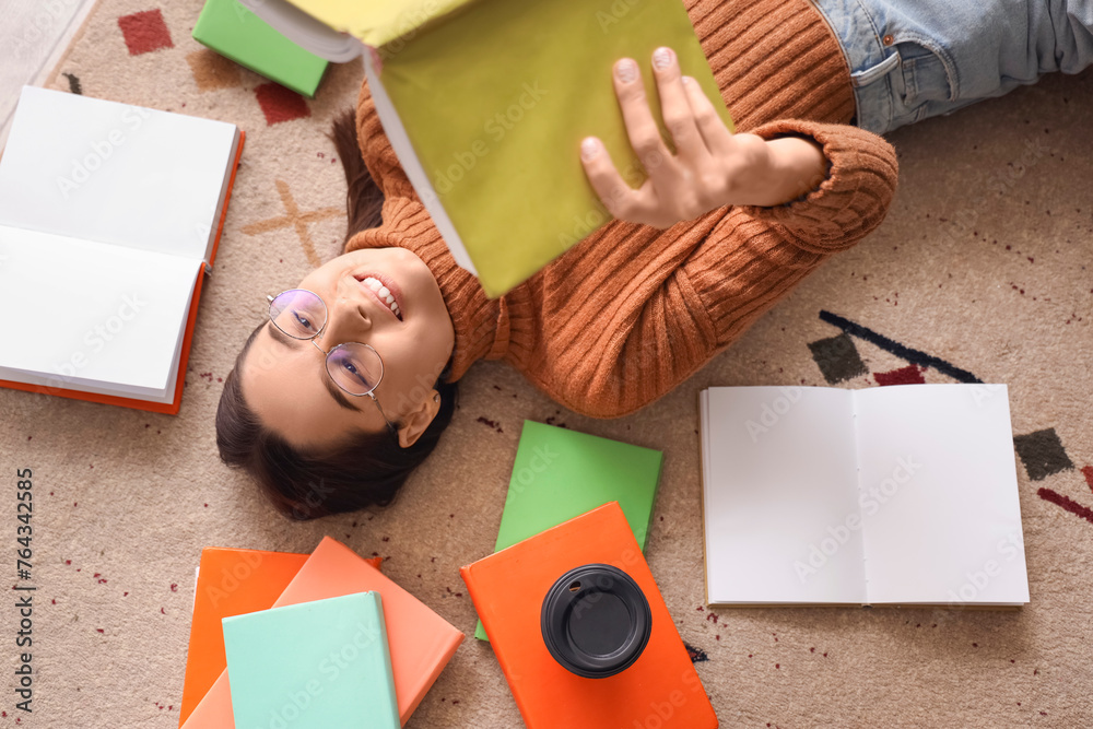 Young woman reading book on carpet at home, top view