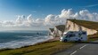 White motorhome parked on a cliffside with blue sky and green grassy hills in the background, overlooking a sea beach with pebbles and waves and rocks.