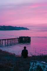 Wall Mural - A man sits on the grassy hill overlooking an old pier that is floating in the ocean, the sky above has pastel pink and purple hues