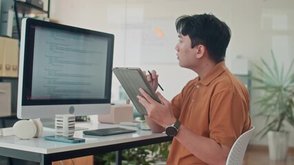 Medium shot of busy Asian developer testing application for computers and making notes on digital tablet