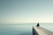 A solitary individual sits at the end of a pier, gazing out at a peaceful sea, lost in contemplation under a clear sky.