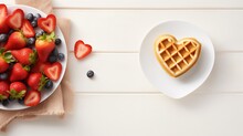 Heart-shaped waffle on a white plate with a bowl of strawberries and blueberries, ideal for a romantic breakfast or Valentine's Day treat.
