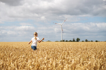 Back view little cute happy blond caucasian girl enjoy walking ripe wheat ear field against wind mill turbine farm on warm sunny summer day. Child freedom future clean energy environment concept