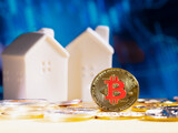 Fototapeta Desenie - Bitcoin coin stands against a blurred blue background with financial graphs and two white model houses in the background. cryptocurrency and real estate. finance and investment on stock market
