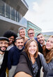 © Gigi Delgado - Vertical image of a cheerful group of multiracial business people taking a selfie together outside office building, looking at the camera smiling. Copy space.