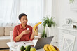 happy african american nutritionist holding apple and giving diet advice on laptop in kitchen