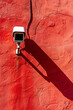 Outdoor security camera, white in color, casting a shadow on a red wall