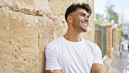 Wall Mural - Handsome young hispanic man smiling outdoors in a sunny urban setting, wearing a casual white shirt and leaning on a stone wall.