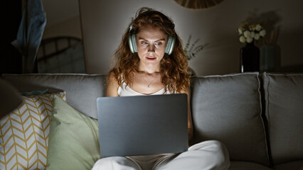 Sticker - A young woman in casual clothing uses a laptop while wearing headphones, sitting on a couch in a cozy living room at night.