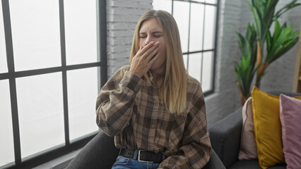 Poster - A tired young woman yawning indoors, sitting comfortably in a cozy, modern apartment living room.