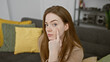 © Krakenimages.com - A young blonde woman in a sweater touches her face thoughtfully inside a cozy living room, reflecting a quiet moment at home.