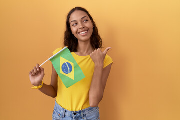 Poster - Young hispanic woman holding brazil flag smiling with happy face looking and pointing to the side with thumb up.