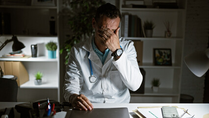 Poster - A stressed man in a white lab coat sitting at a desk in a hospital room, portraying exhaustion or concern.