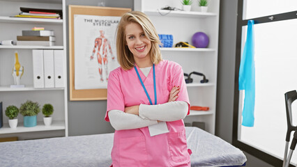 Confident young woman healthcare professional with short blonde hair, smiling in a pink scrub with crossed arms in a modern clinic interior.