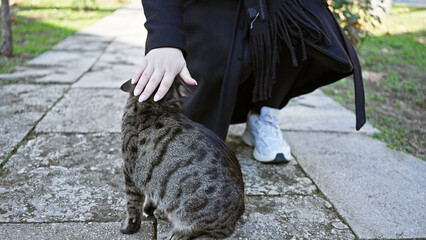 adult woman in casual attire petting a tabby cat on a paved path in an urban park setting.