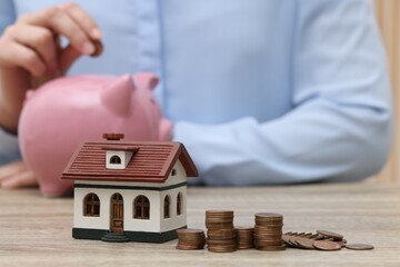 Wall Mural - Woman with piggy bank at wooden table, focus on house model and stacked coins