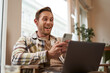 Image of man in cafe, coffee shop visitor sitting in chair with laptop and smartphone, looking surprised and excited at mobile phone screen, amazed by big news, promo offer on app