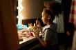© Seventyfour - Side view portrait of little Black girl sitting at vanity mirror backstage and doing makeup preparing for theater performance