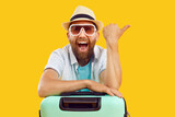 Fototapeta Natura - Portrait of young happy funny man tourist in sunglasses and beach hat with suitcase pointing index finger to the side on yellow background. Vacation advertising, summer holiday and travel concept.