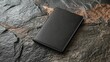 Black leather book on a stone background. The book is closed and the spine is facing the camera. The leather is smooth and has a slight sheen.