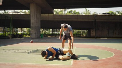 Wall Mural - 4K Caucasian man basketball player lifting fallen friend on outdoors court. Sportsman do sport training streetball at street court under city highway. Fair game competition and sportsmanship concept.