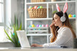 Side view portrait of businesswoman sitting at the home office desk and looking at laptop screen. She is wearing bunny ears headphones. Colorful easter eggs in the background.