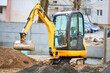© R_Yosha - Tracked orange mini excavator digger near road with green trees and concrete fence on background with copyspace.