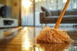 Sunlight floods a clean living room as an orange mop rests on the glossy wooden floor, implying a sense of comfort and tidiness