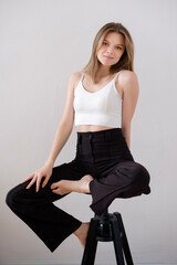 a young pretty brown-haired girl with daytime light makeup and well-groomed hair in a white top and black pants sits barefoot on a bar stool against a white wall