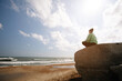 A young woman sits on a stone and relaxes or does yoga, meditation. On the background there is sea and sky, sun rays. She sits with her back turned.