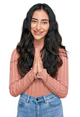 Hispanic teenager girl with dental braces wearing casual clothes praying with hands together asking for forgiveness smiling confident.