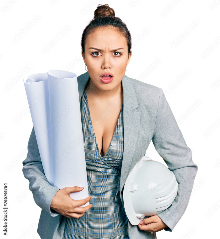 Young hispanic girl holding safety helmet and blueprints afraid and shocked with surprise and amazed expression, fear and excited face.