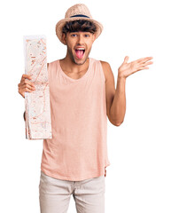 Wall Mural - Young hispanic man wearing summer hat holding map celebrating victory with happy smile and winner expression with raised hands